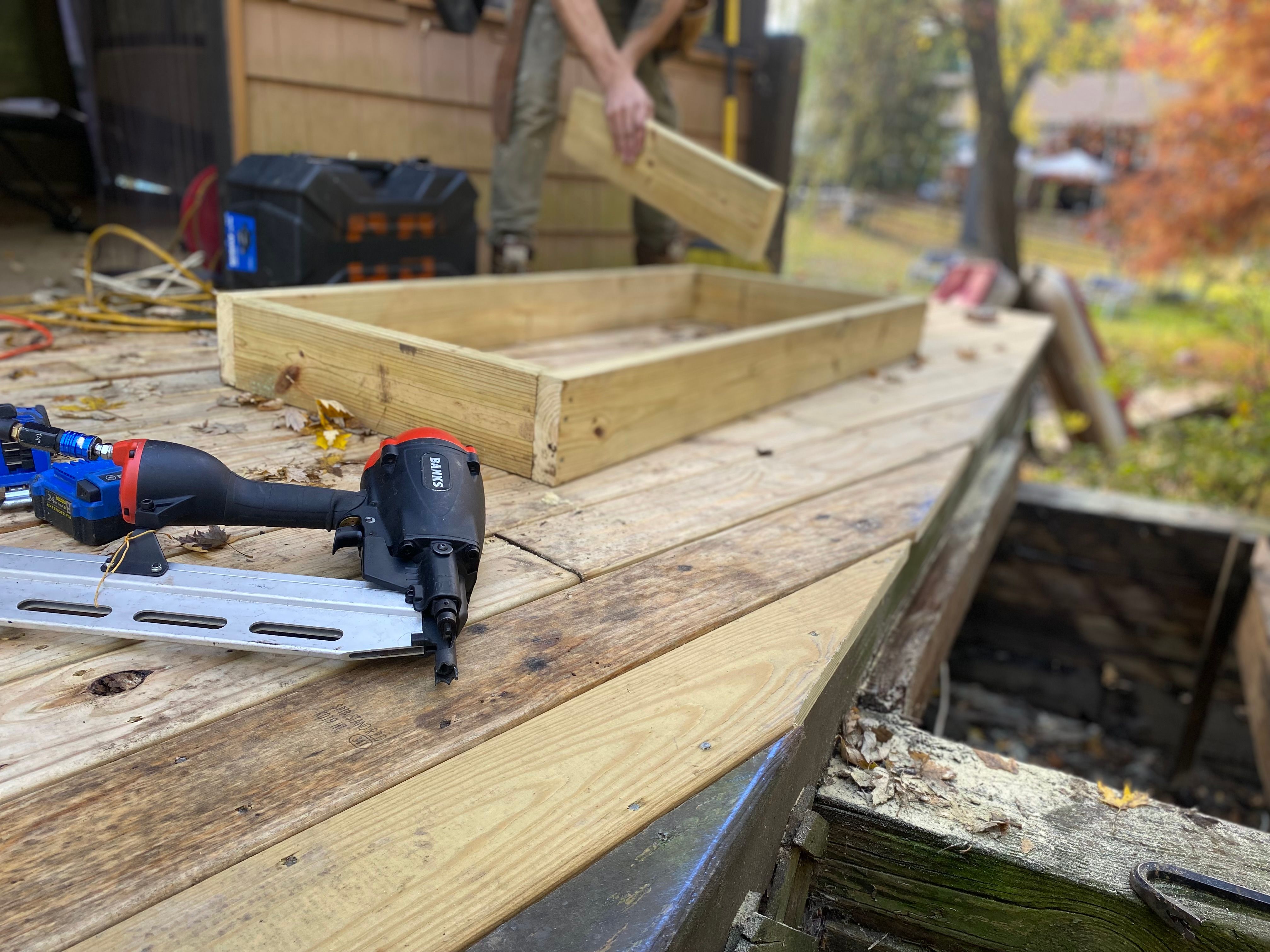 Close-up of deck construction with a worker using a nail gun on a framed outdoor floor.