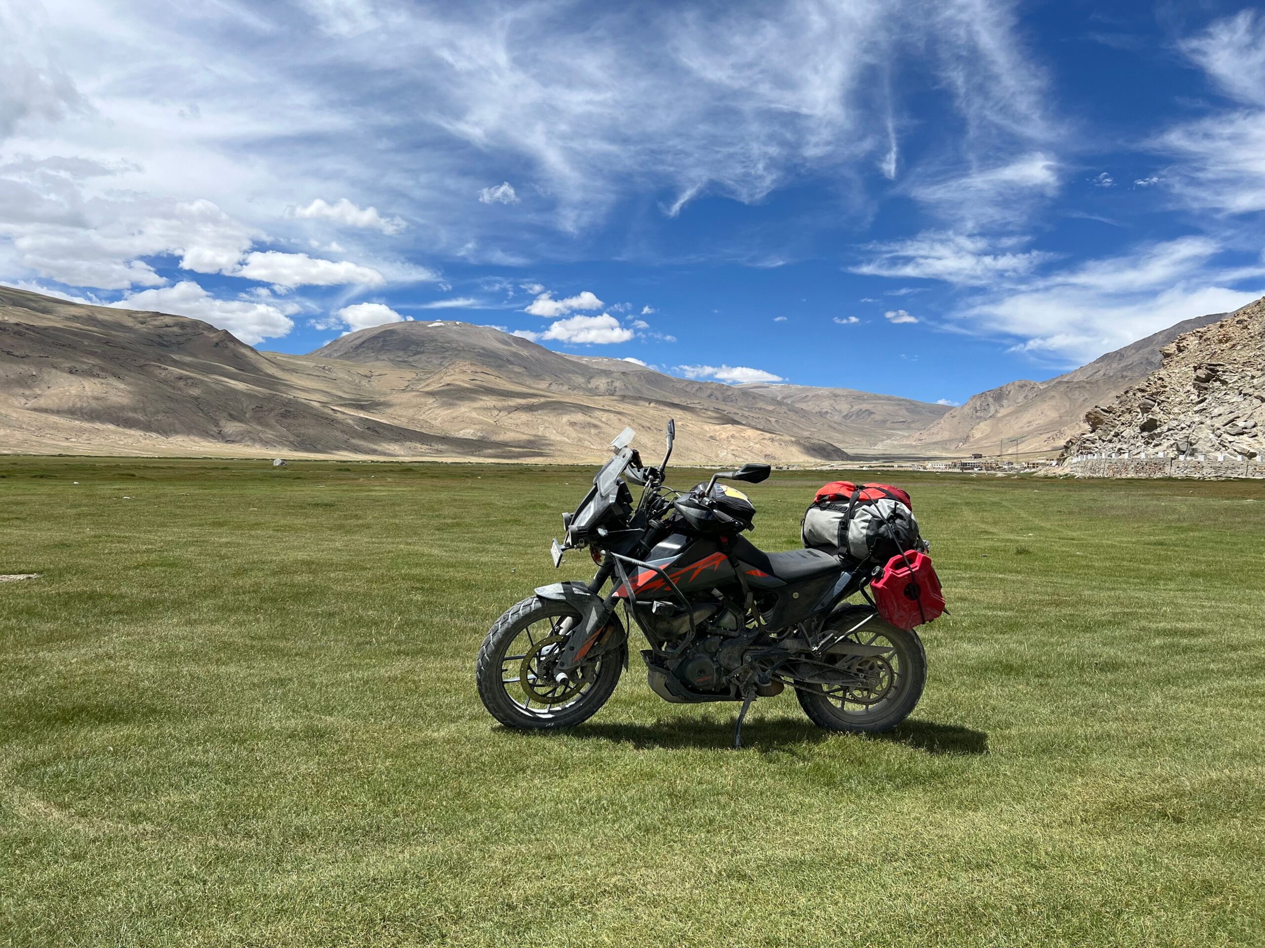 A motorcycle loaded with travel gear stands in a broad green landscape with mountains in the distance.